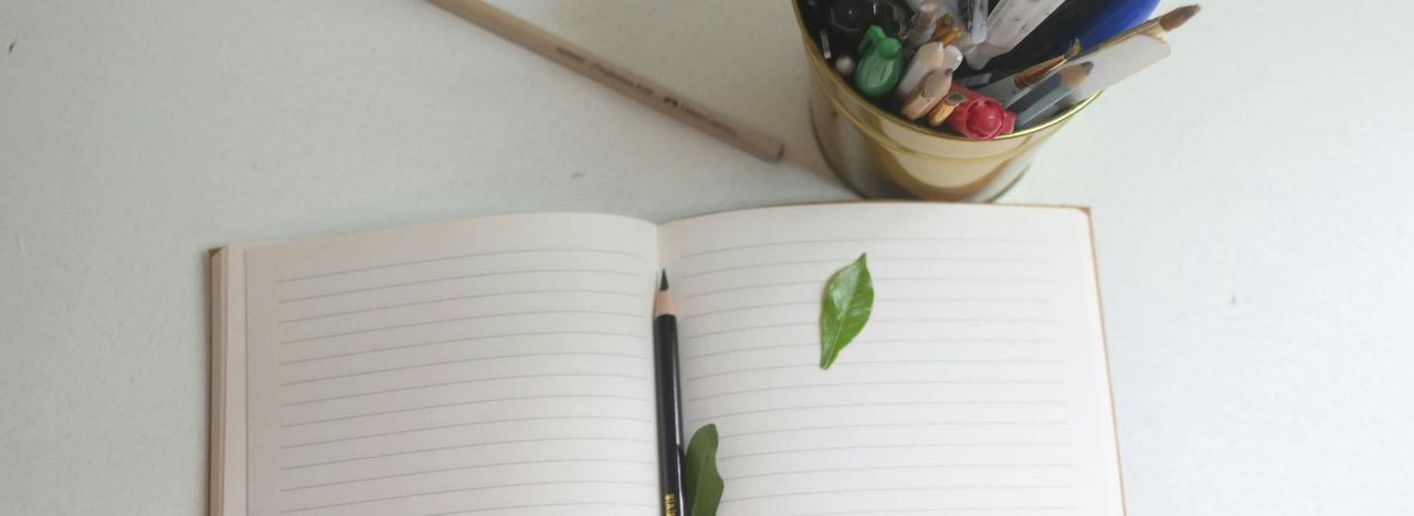 An open and blank notebook with a couple of leaves, a stationery holder cup with pens, all on a white table, representing Arts and Trauma healing course at All Nations.