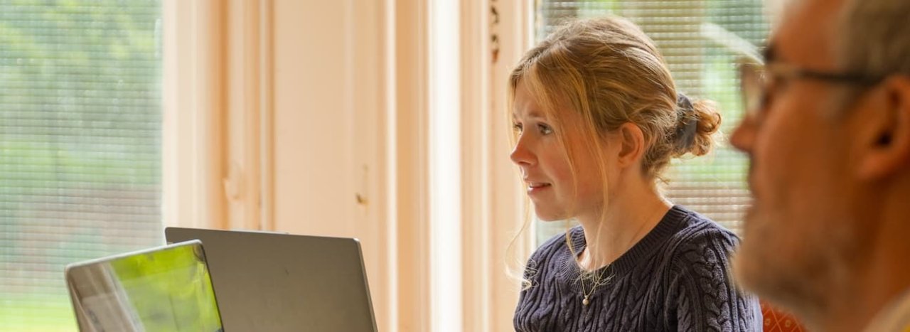 A young lady in a blue top, intent in study and listening, next to an older man out of focus, next to 2 laptop screens, representing the Contextual Theology Contemporary issues.