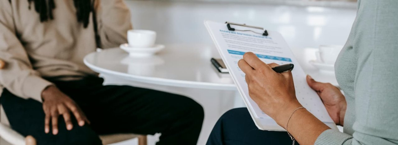 a man and a professional woman sat at a white table with a white coffee cup while the professional fills in a form by pen, representing the course on  staff care wellbeing and christian mission.