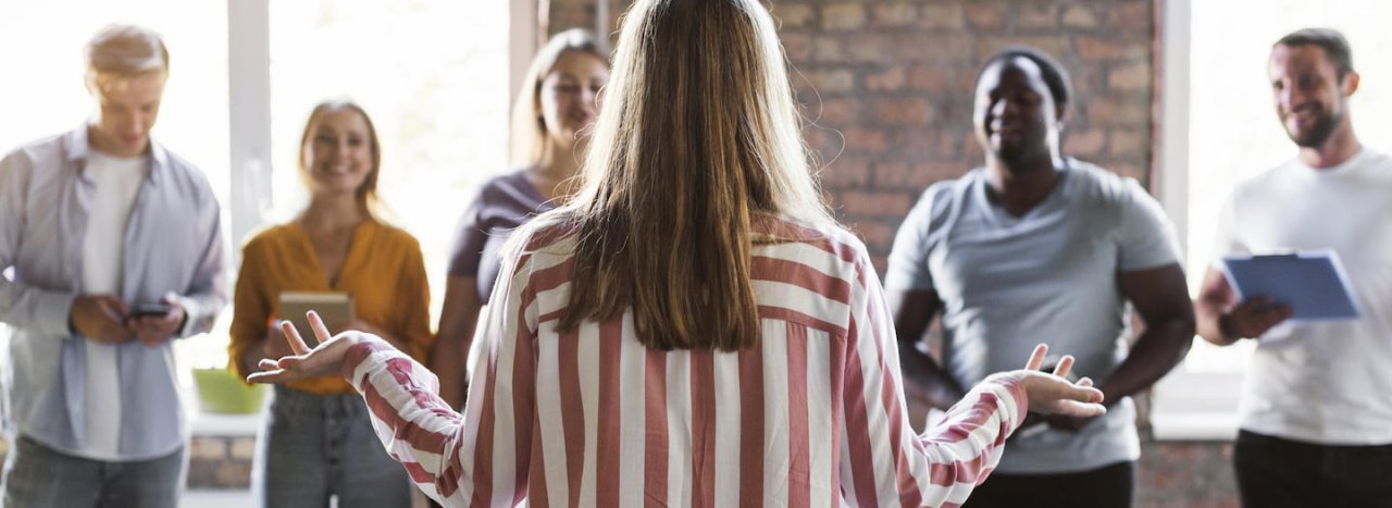 A female leader standing with her back to the viewer, addressing a group of younger people of different backgrounds, representing the intercultural leadership courses at All Nations college.