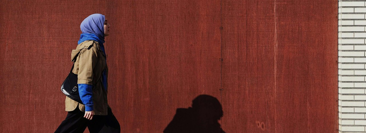 A muslim veiled woman walking past a wooden wall with a white brick ending, showing that our mission if everyone from all walks of life.
