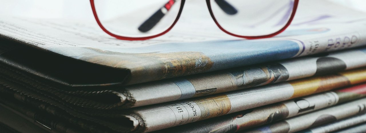 A stack of newspapers folded with a pair of reading glasses propped above, signifying the news page for All Nations.
