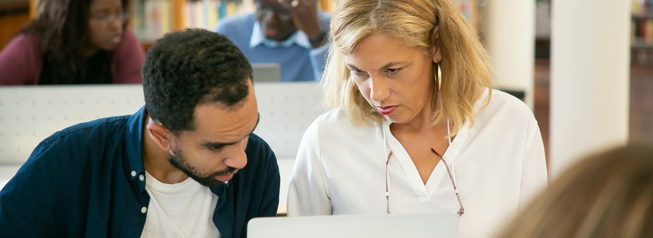 An older lady supporting and helping a younger learner or person in front of a laptop with other people (various ethnicities) in the background.