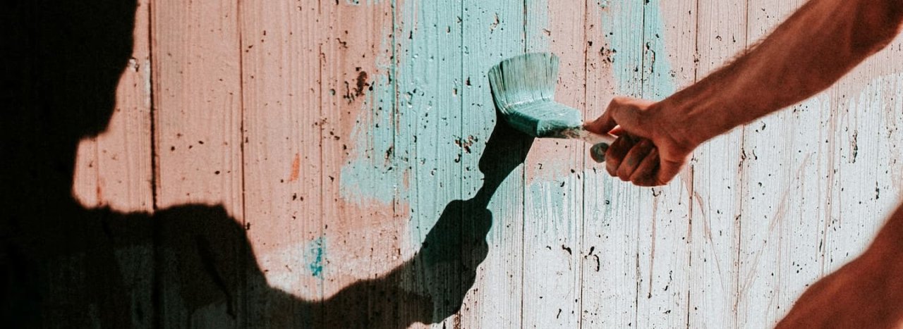 A man's arm painting a fence from mauve to pastel blue, with a strong silhouette reflection, representing volunteering across all areas at All Nations.
