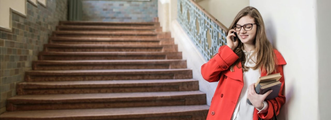 A young woman or student holding some books in a more traditional staircase and holding a phone, representing contacting All Nations.