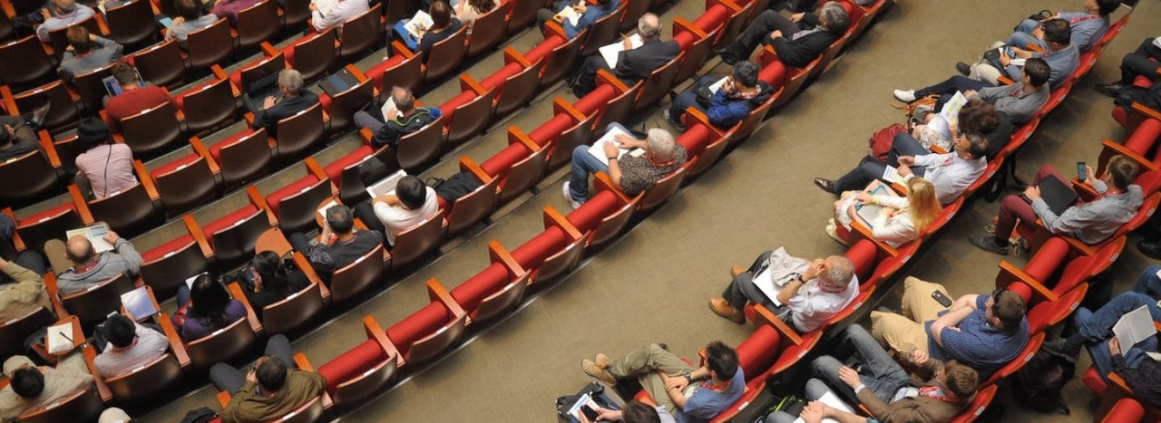 Rows of red chairs in an auditorium with various people sitting in them, representing a conference which All Nations might attend.
