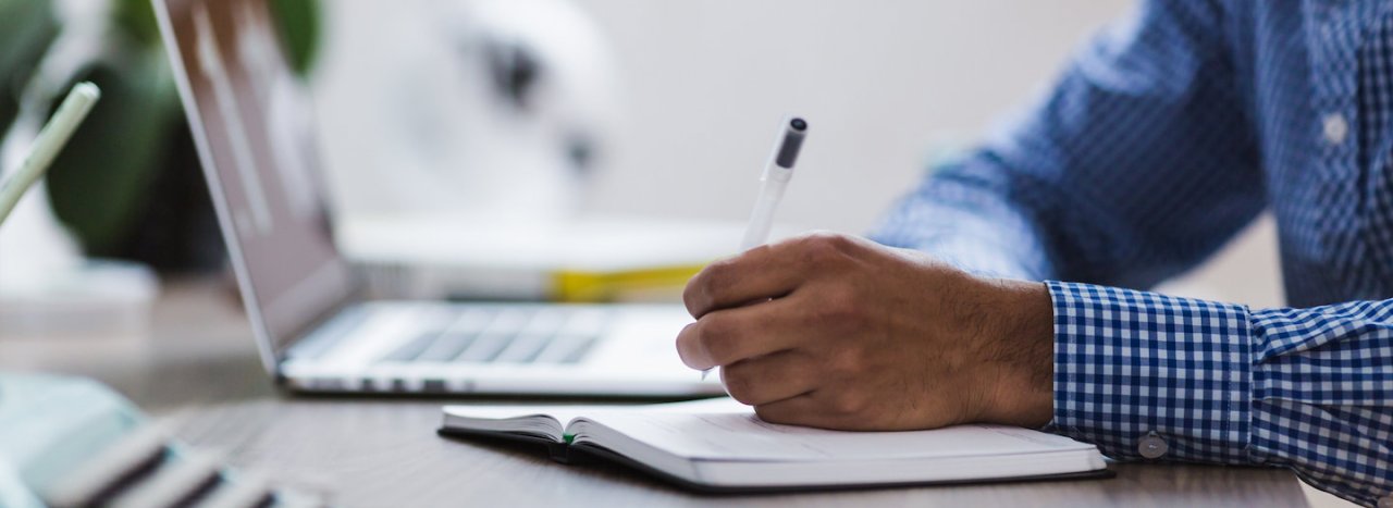 A man wearing a blue and white shirt, holding a pen on a notebook, near a laptop, representing the application process to All Nations college.