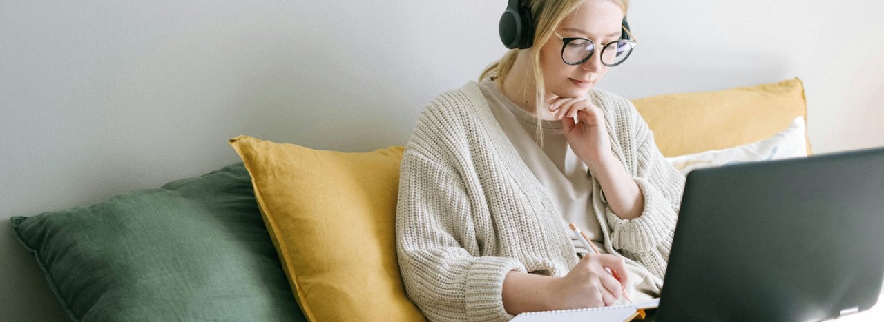 A young student dressed in light colours with headphones and glasses busy studying at a laptop and notebook, representing flexible and open learning from a distance, typical of the open university.