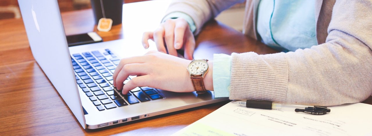 An older student dressed more traditionally, working at a laptop with a book and pen beside him and a phone and cup, representing postgraduate course application at All Nations college.