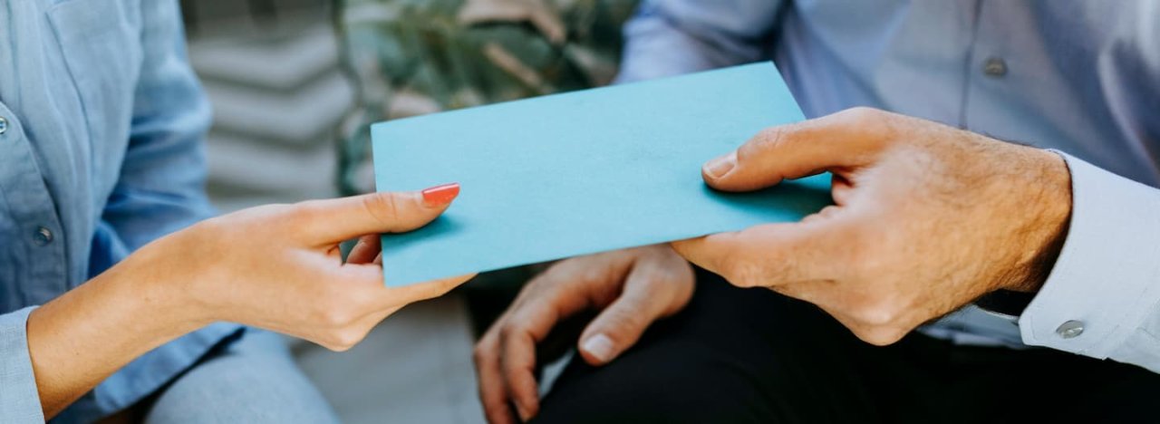 A blue envelope with money being gifted from one person to another, representing financial, monetary and prayer giving to All Nations.